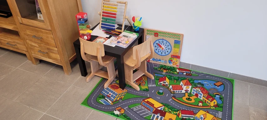 Spielbereich mit einem Tisch, Stühlen, Bausteinen und einem Teppich mit Straßenmuster.